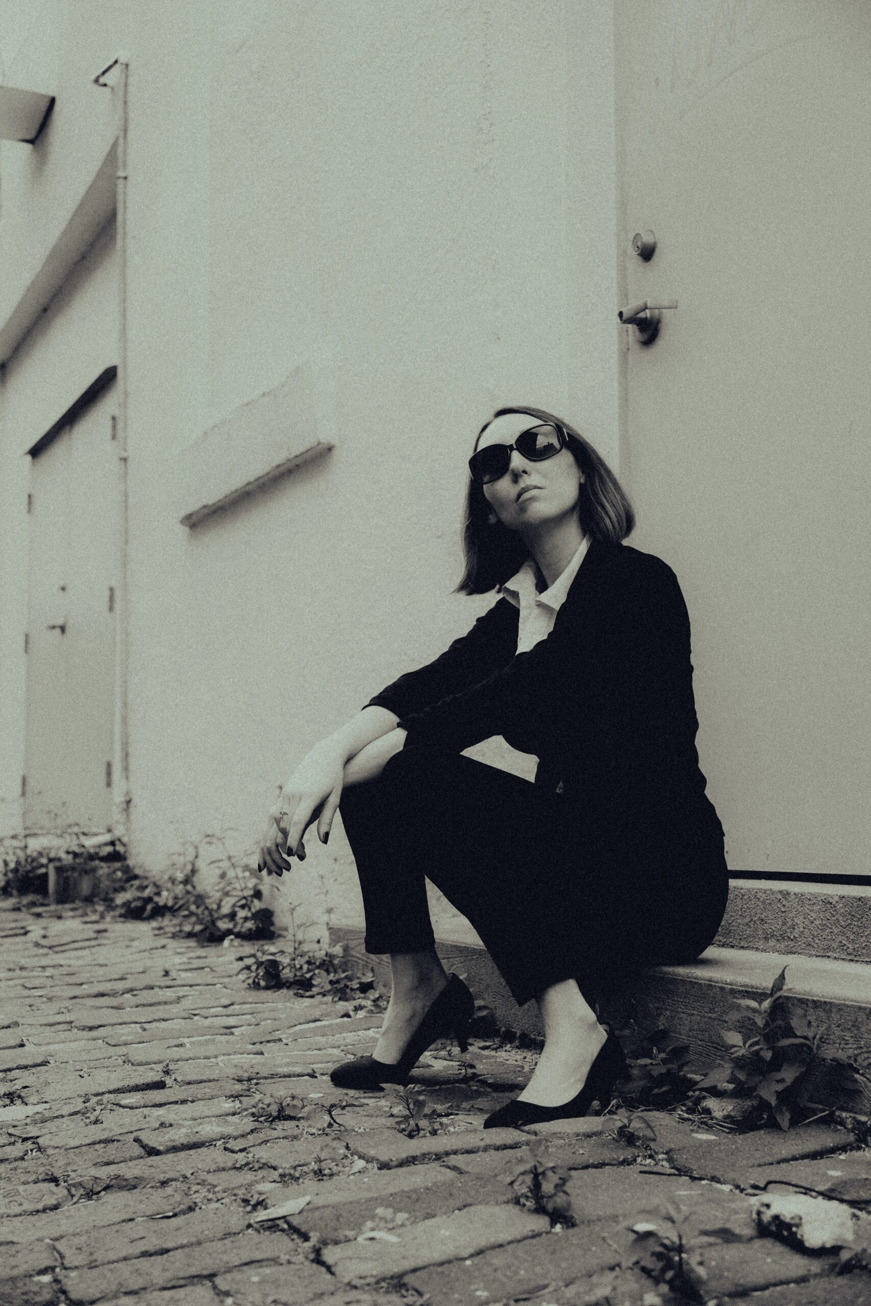 A woman wearing dark sunglasses, a business suit, and high heels sits outside on a concrete step.